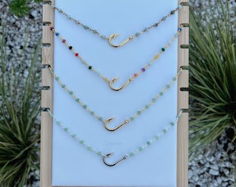 May include: Four gold-colored necklaces with fish hook pendants. Each necklace features a different colored bead strand. The necklaces are displayed on a white background with a wooden frame, showcasing the jewelry's design and colors.
