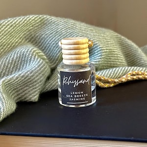 May include: A small clear glass bottle with a ribbed wooden cap and a black label featuring the text Rhysand, Lemon, Sea Breeze, and Jasmine. The bottle has a gold and white string attached to the top, designed for hanging as an air freshener.