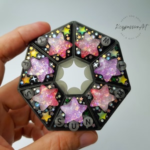 Puede incluir: Un organizador de pastillas para siete días con fondo negro y diseño de estrellas. Cada compartimento tiene una estrella de color diferente con purpurina y una etiqueta del día de la semana: Dom, Lun, Mar, Mié, Jue, Vie, Sáb.