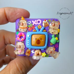 May include: A purple square-shaped resin charm with a cartoon television, donuts, hearts, and bears. The charm is decorated with colorful details and has a glossy finish.