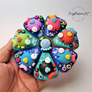 Puede incluir: Un colorido juguete artesanal con temática de peces y una figura central de pulpo. El juguete presenta seis segmentos, cada uno con un diseño de pez diferente en varios colores y letras que deletrean los días de la semana. El juguete se sostiene en una mano.