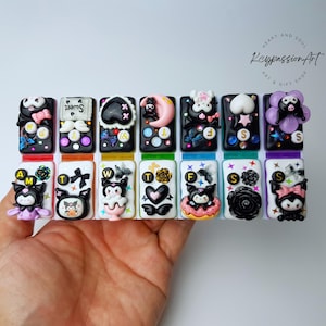 May include: A set of 10 colorful keychains featuring a black and white cartoon character with a bow. Each keychain has a letter of the alphabet and a different design. The keychains are arranged in a row on a hand.