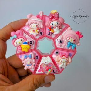 May include: A pink and white pill organizer shaped like a flower, featuring cartoon characters with bows. Each compartment is labeled with a letter. The organizer is held in a hand, with the text "Heart and Soul Keypassion Art & Gift Shop" visible.