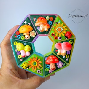 Op de afbeelding: Een kleurrijk, geometrisch speelgoed met paddenstoel- en bloemontwerpen. Elke sectie toont een dag van de week, met de letters gerangschikt rond de decoratieve elementen. Het speelgoed wordt in een hand gehouden.