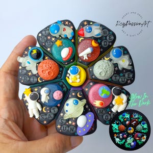 Puede incluir: Un juguete fidget negro de varias secciones con temática espacial con varios planetas, astronautas y un cohete. El juguete brilla en la oscuridad e incluye el texto "Glow In The Dark". El diseño incorpora el texto "KeyPassionArt".