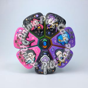 May include: A circular, flower-shaped pill organizer with six wedge-shaped compartments. Each section features unique anime-style character charms, glittery accents, and letters representing days of the week. The center has a small, oval blue gem.