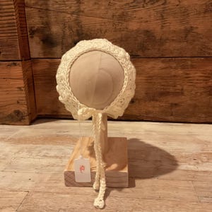 Puede incluir: Un gorro de bebé de ganchillo blanco con un lazo atado. El gorro se muestra en un soporte de madera.