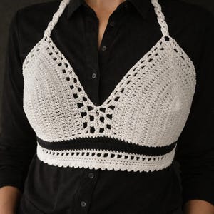 May include: A white crochet halter top with a black band at the bottom. The top is made of a textured, open-weave crochet pattern. The halter straps are also crocheted. The top is worn over a black button-down shirt.