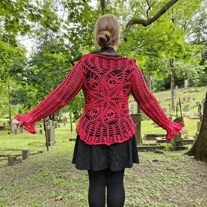 May include: A red crochet top with a skull design, long sleeves, and flared cuffs. The top is worn over a black outfit. The background features a cemetery with green grass and trees.
