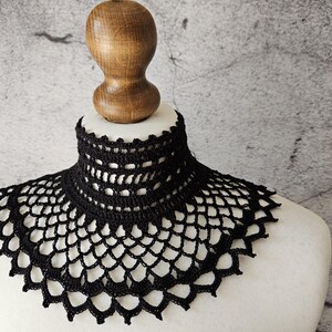 May include: A black, handmade crochet collar displayed on a mannequin. The collar features a high, close-fitting neck and a scalloped edge. The openwork design is visible, showcasing the intricate craftsmanship of the piece.
