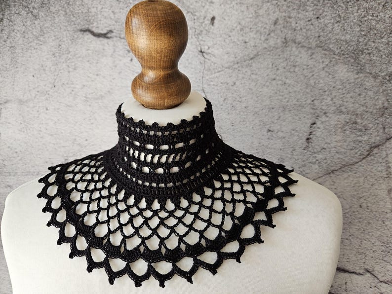 May include: A black, handmade crochet collar displayed on a mannequin. The collar features a high, close-fitting neck and a scalloped edge. The openwork design is visible, showcasing the intricate craftsmanship of the piece.