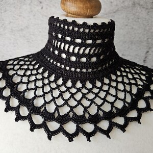 May include: A black crochet collar with a high neck and a scalloped edge. The collar is made of a delicate, open-weave pattern, creating a lace-like appearance. The collar is displayed on a white mannequin.