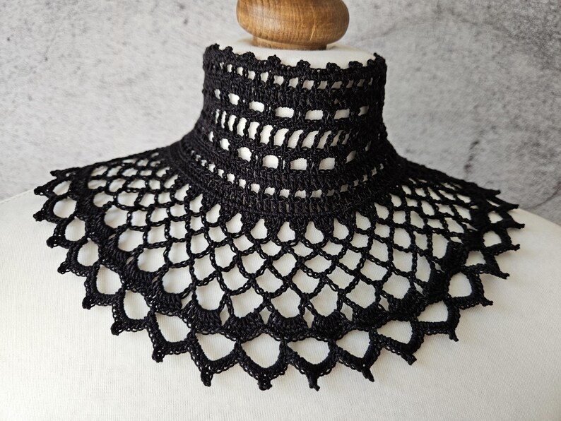 May include: A black crochet collar with a high neck and a scalloped edge. The collar is made of a delicate, open-weave pattern, creating a lace-like appearance. The collar is displayed on a white mannequin.