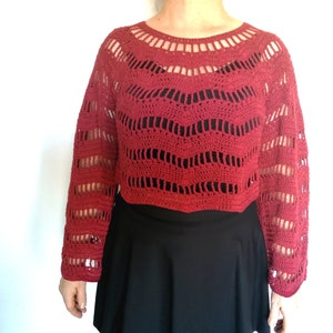 Puede incluir: Un top de crochet rojo con mangas largas y un ajuste holgado. El top tiene un escote amplio y abierto y un delicado patr&oacute;n de encaje.