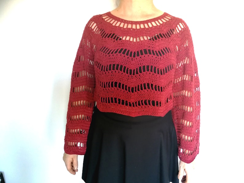 Puede incluir: Un top de crochet rojo con mangas largas y un ajuste holgado. El top tiene un escote amplio y abierto y un delicado patr&oacute;n de encaje.