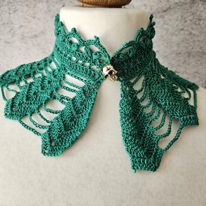 May include: A teal green crocheted collar with an openwork design. The collar is fastened with a gold-colored skull clasp. The collar is displayed on a white mannequin head.