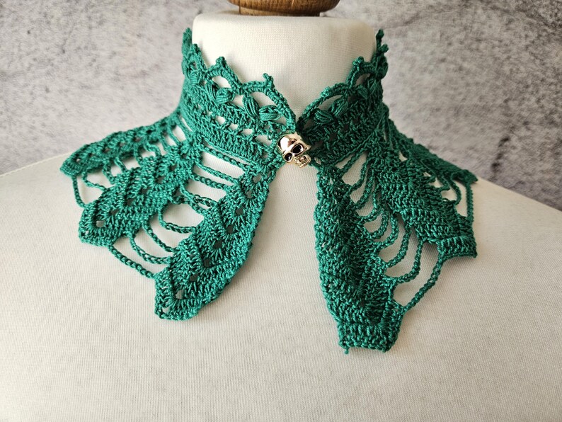 May include: A teal green crocheted collar with an openwork design. The collar is fastened with a gold-colored skull clasp. The collar is displayed on a white mannequin head.