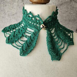 May include: A handmade, emerald green crochet collar with a decorative skull clasp. The collar is displayed on a white mannequin, showcasing the intricate openwork design and the detailed skull clasp.