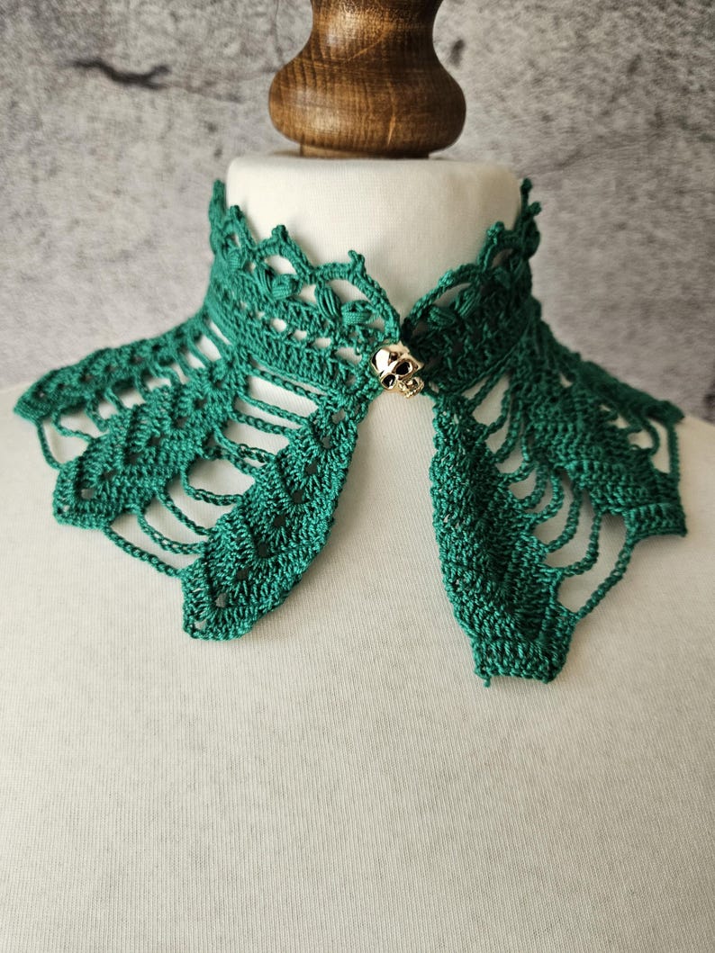 May include: A handmade, emerald green crochet collar with a decorative skull clasp. The collar is displayed on a white mannequin, showcasing the intricate openwork design and the detailed skull clasp.