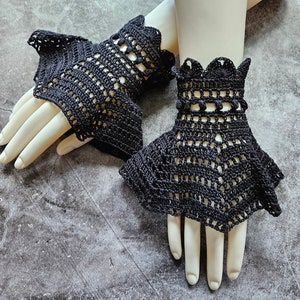 Puede incluir: Un par de puños de muñeca de crochet negro con un diseño de encaje y pequeñas bolitas. Los puños se muestran en una mano de maniquí.