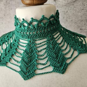 May include: A handmade, emerald green crochet collar. The collar features a detailed openwork design with a scalloped edge and a geometric pattern. The collar is displayed on a white mannequin head, showcasing its intricate craftsmanship.