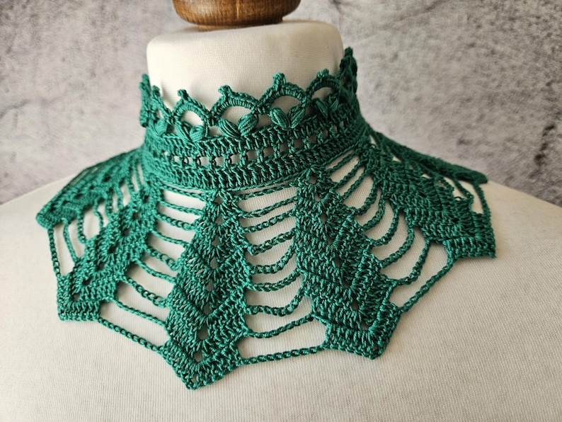 May include: A handmade, emerald green crochet collar. The collar features a detailed openwork design with a scalloped edge and a geometric pattern. The collar is displayed on a white mannequin head, showcasing its intricate craftsmanship.