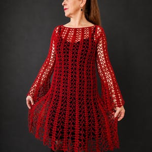 May include: A red crochet dress with long sleeves and a flared skirt. The dress features an open, lacy design, revealing a black underdress. The model is standing against a dark grey background.