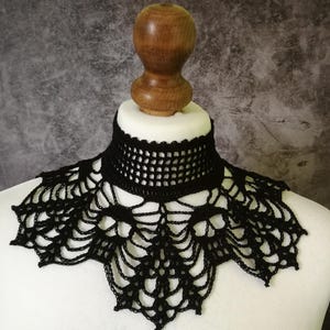 Puede incluir: Cuello de ganchillo negro con dise&ntilde;o de telara&ntilde;a. El cuello se muestra en un busto de maniqu&iacute; blanco con cuello de madera. El cuello tiene un escote tipo gargantilla y un patr&oacute;n calado detallado.
