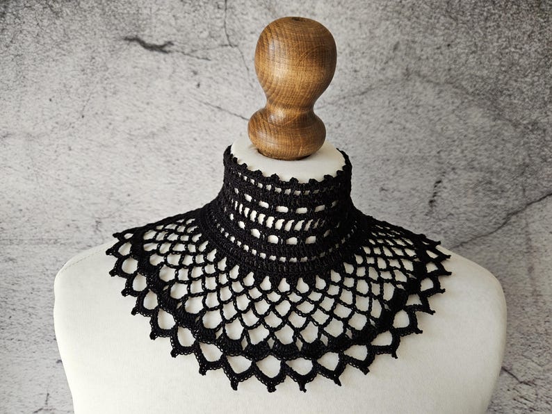 May include: A black crocheted collar with a scalloped edge and openwork design. The collar is displayed on a white mannequin bust with a wooden neck piece, set against a textured gray background. The collar is a fashion accessory.