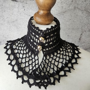 May include: Black crocheted collar with a scalloped edge and skull-shaped buttons. The collar is displayed on a wooden mannequin head. The openwork design and skull accents give the collar a gothic aesthetic.