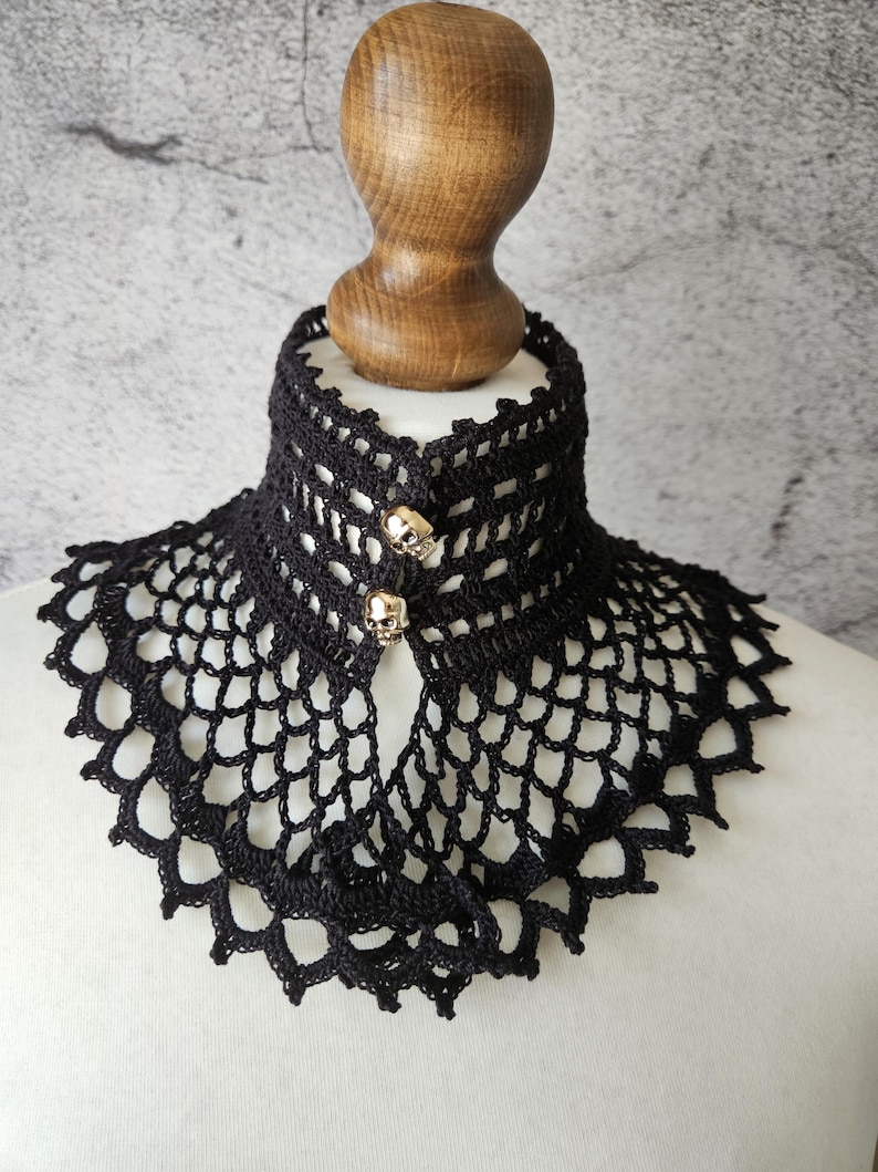 May include: Black crocheted collar with a scalloped edge and skull-shaped buttons. The collar is displayed on a wooden mannequin head. The openwork design and skull accents give the collar a gothic aesthetic.