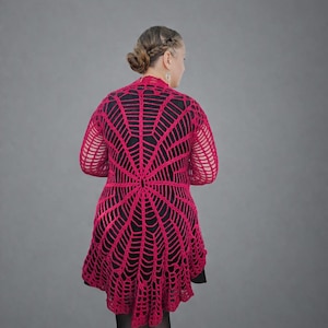 Peut inclure: Un cardigan en crochet fuchsia vif avec un motif de toile d'araignée. Le motif ajouré crée un aspect délicat et aéré. Le cardigan arrive au genou et a des manches longues. Le modèle porte un haut noir en dessous, créant un contraste avec le rose vif.