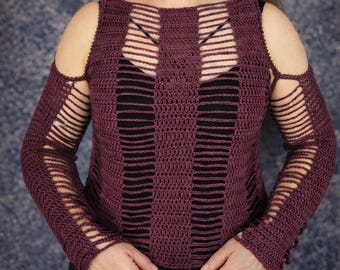 May include: A plum-colored crochet top with long sleeves and a round neckline. The top features vertical and horizontal cut-out details across the chest and sleeves. The sleeves have cut-out slits along the upper arm. The top is worn over a black garment.