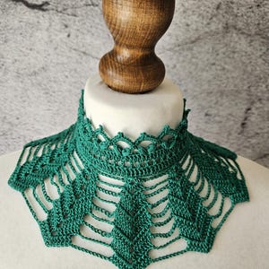 May include: A handmade, emerald green crochet collar with a delicate openwork design. The collar is displayed on a white mannequin head, showcasing its intricate details and texture. The collar's design features a scalloped edge and geometric patterns.