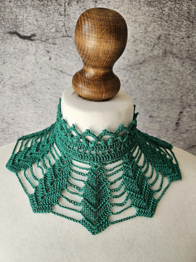 May include: A handmade, emerald green crochet collar with a delicate openwork design. The collar is displayed on a white mannequin head, showcasing its intricate details and texture. The collar's design features a scalloped edge and geometric patterns.