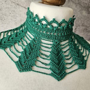 May include: A handmade, emerald green crochet collar. The collar features a high, detailed neckline with a scalloped edge and a delicate, openwork design that drapes down. The intricate pattern and rich color create a unique accessory.