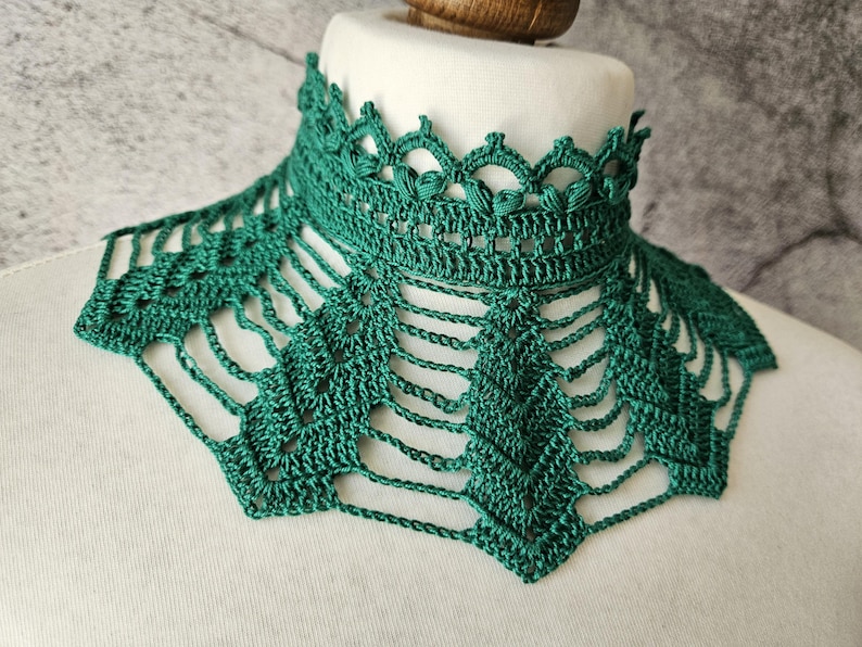 May include: A handmade, emerald green crochet collar. The collar features a high, detailed neckline with a scalloped edge and a delicate, openwork design that drapes down. The intricate pattern and rich color create a unique accessory.