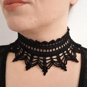 Może przedstawiać: Czarny szydełkowy naszyjnik typu choker z misternym ażurowym wzorem. Naszyjnik ma falisty brzeg i delikatny, koronkowy wygląd. Choker jest noszony wokół szyi i jest stylowym dodatkiem.