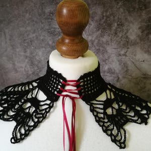 Puede incluir: Cuello de ganchillo negro con cierre de lazo de cinta roja. El cuello se muestra en un busto de maniqu&iacute; blanco. El dise&ntilde;o de encaje y la cinta roja a&ntilde;aden un toque de elegancia a este accesorio &uacute;nico.