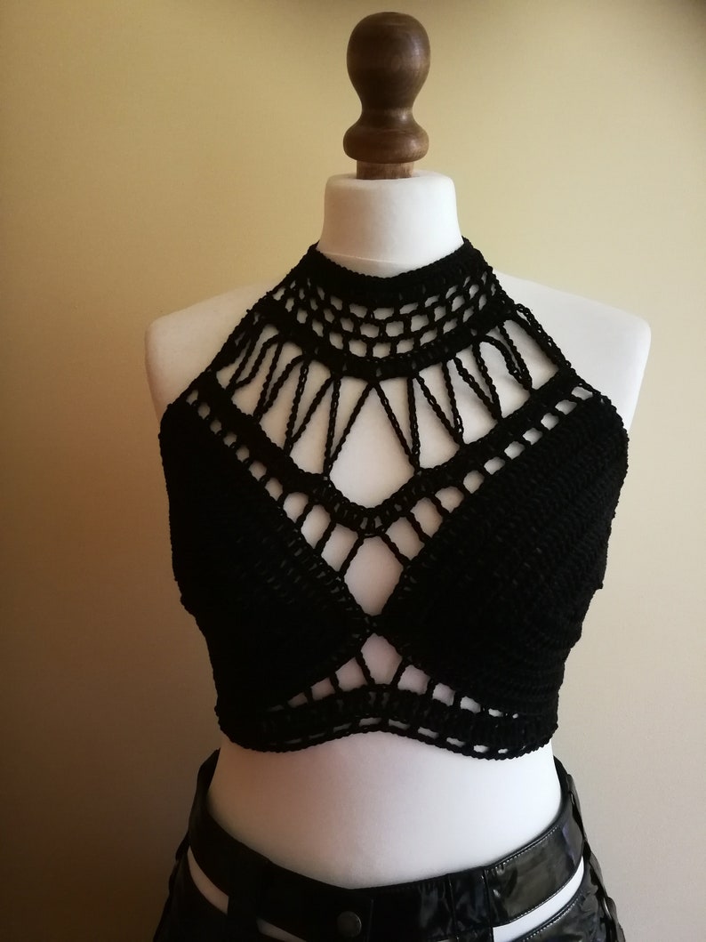 May include: A black crochet halter top displayed on a mannequin. The top features a high neckline and a geometric design across the chest. The crochet work is intricate, with open spaces creating a textured look. The top is a solid black color.