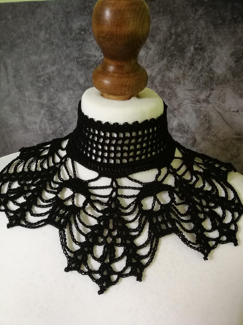 Puede incluir: Un collar negro de ganchillo con un dise&ntilde;o similar a una telara&ntilde;a. El collar tiene un escote alto y estructurado y un borde festoneado. El intrincado encaje se muestra sobre una cabeza de maniqu&iacute; blanca sobre un fondo gris.
