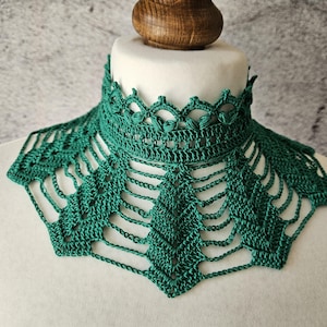 May include: A handmade, emerald green crochet collar. The collar features a high, detailed neckline with a scalloped edge and a geometric openwork design. The collar is displayed on a white mannequin bust against a gray background.