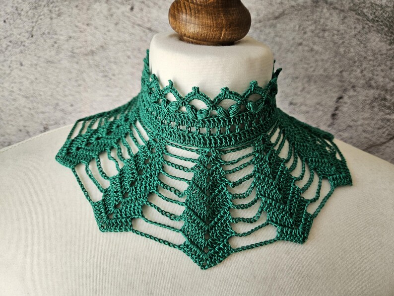 May include: A handmade, emerald green crochet collar. The collar features a high, detailed neckline with a scalloped edge and a geometric openwork design. The collar is displayed on a white mannequin bust against a gray background.