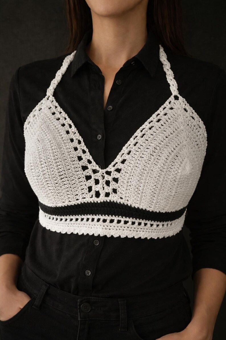 May include: A white crochet halter top with a black band at the bottom. The top is made of a textured, open-weave crochet pattern. The halter straps are also crocheted. The top is worn over a black button-down shirt.