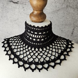 May include: A black crocheted collar with a high neckline and a scalloped edge. The collar is displayed on a white mannequin bust with a wooden neck and head. The intricate openwork design is visible.