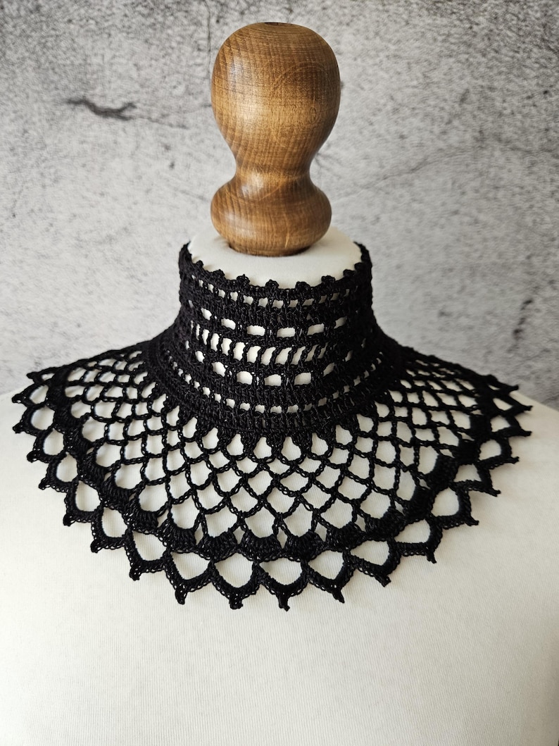 May include: A black crocheted collar with a high neckline and a scalloped edge. The collar is displayed on a white mannequin bust with a wooden neck and head. The intricate openwork design is visible.