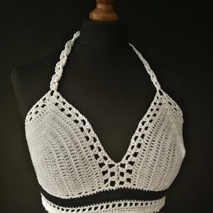 May include: White crochet halter top with a black stripe detail. The top features a V-neck design and a scalloped edge. The halter straps are also crocheted. The top is displayed on a black mannequin.