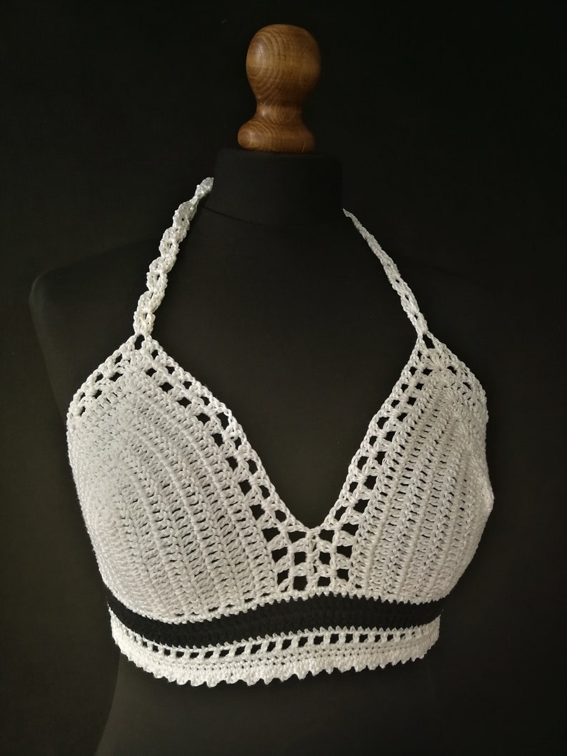 May include: White crochet halter top with a black stripe detail. The top features a V-neck design and a scalloped edge. The halter straps are also crocheted. The top is displayed on a black mannequin.