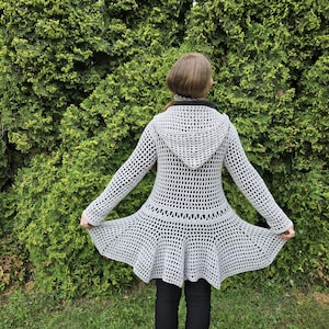 May include: A light gray, open-weave crochet coat with a hood and flared skirt. The long-sleeved coat is knee-length and worn over black pants. The background is a dense green hedge.
