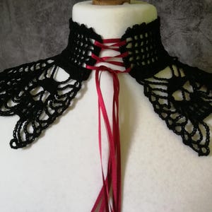 Puede incluir: Un cuello negro de ganchillo con un delicado dise&ntilde;o calado. El cuello presenta una cinta roja que se entrelaza en la parte posterior, creando un detalle tipo cors&eacute;. El cuello se muestra sobre un busto de maniqu&iacute; blanco.